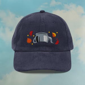 May include: Navy blue corduroy baseball cap with an embroidered design. The design features a gray teapot with a white handle and spout, along with embroidered autumn leaves in red, orange, and yellow.