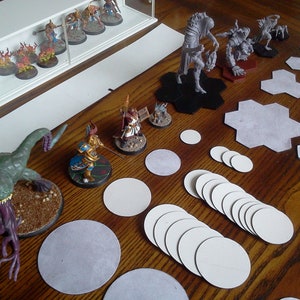 May include: A collection of miniature fantasy figures, including warriors, monsters, and creatures, arranged on a wooden surface. Some figures are displayed in a clear case. Also includes various game pieces, such as hexagonal and circular tiles.