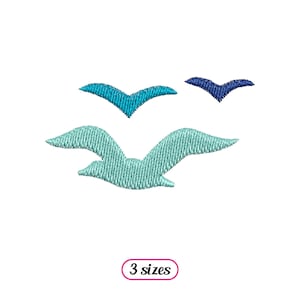 Puede incluir: Tres gaviotas bordadas en tonos de azul y verde. La gaviota más grande está en primer plano y es de color verde claro. Las dos gaviotas más pequeñas están en el fondo y son de color azul oscuro.