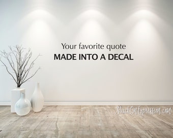 Op de afbeelding: Een witte muur met de tekst "Your favorite quote MADE INTO A DECAL" in zwarte letters. De muur bevindt zich in een kamer met een witte vloer en drie witte vazen met takken.