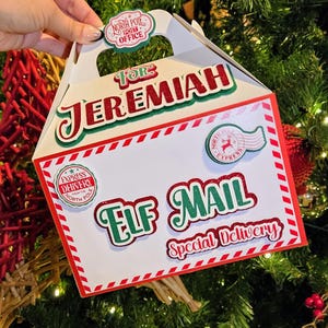 May include: A white Christmas-themed gift box with red and green accents. The box features the text "FOR JEREMIAH" and "ELF MAIL Special Delivery". Decorative stickers include "North Pole Post Office" and "Express Delivery".