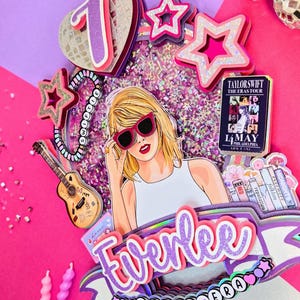 May include: A colorful cake topper with a Taylor Swift theme. It features a cartoon illustration of Taylor Swift wearing sunglasses, surrounded by stars, a guitar, and the text "Everlee". The topper includes the text "7 YEAR ERA" and a concert date.