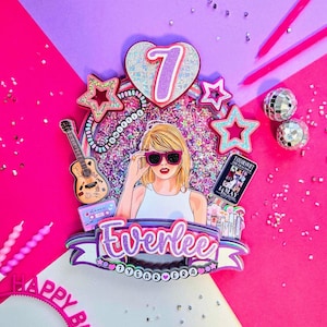 May include: A colorful birthday cake topper featuring a cartoon illustration of a woman wearing sunglasses. The topper includes the name "Everlee", the number "7", stars, a guitar, and a book. The background is pink and purple.