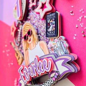 May include: A layered cake topper featuring a cartoon illustration of a blonde woman wearing pink sunglasses. The topper includes the text "Everlee" in purple, a concert poster, and star-shaped decorations. The color scheme is primarily pink and purple.