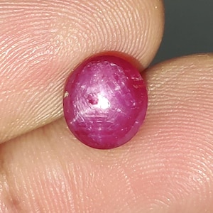 Può includere: Una gemma di rubino lucidata, di forma ovale. La gemma è di un colore rosso intenso e saturo, con una leggera sfumatura violacea. La superficie del rubino ha una qualità liscia e riflettente, con strutture interne visibili. La gemma è tenuta tra due dita.