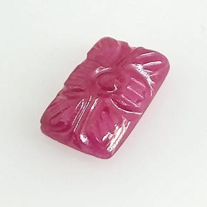 May include: A rectangular, deep pink gemstone featuring a carved floral pattern on its surface. The stone has a polished, glossy finish with distinct, recessed lines forming the petals of a flower design centered on the top face of the gem.