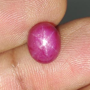 May include: A vibrant, oval-shaped cabochon-cut star ruby gemstone with a deep magenta-pink hue. The stone displays a distinct six-rayed star effect across its smooth, polished surface, reflecting light from a central point within the gemstone.