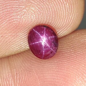 May include: A deep magenta star ruby gemstone with a smooth, polished cabochon cut. The stone displays a distinct six-rayed star effect, known as asterism, which radiates from the centre of the oval-shaped surface when illuminated by light.
