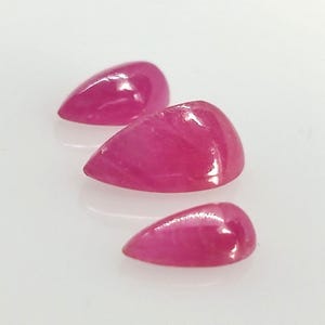 May include: Three polished, teardrop-shaped gemstones in varying sizes. The gems are a vibrant, translucent pink-red color, with a smooth, reflective surface. The stones are arranged on a white surface, with soft shadows.