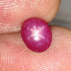 May include: A polished, oval-shaped cabochon gemstone with a deep, vibrant magenta-red hue. The surface displays a distinct six-rayed star effect, known as asterism, which appears as a bright, centered light reflection against the translucent stone.