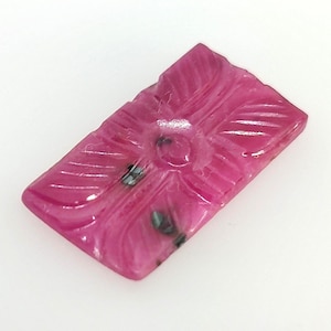 May include: A rectangular, carved gemstone in a vibrant magenta hue. The stone features a central floral design with radiating leaf-like patterns. Small, dark inclusions add subtle contrast to the polished surface. This gemstone is ideal for jewelry making.