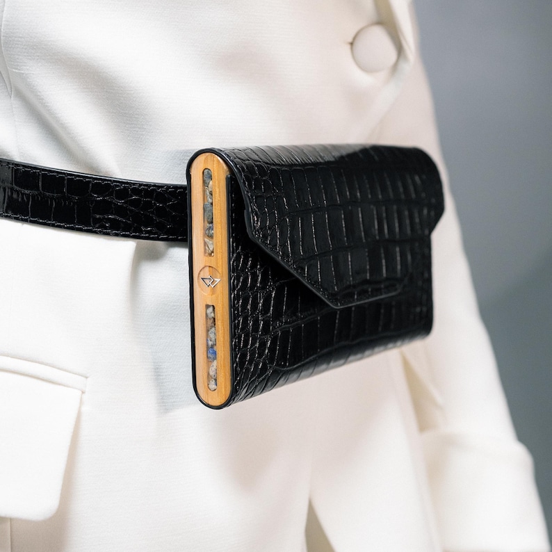 May include: A black crocodile-embossed leather fanny pack with a wooden frame and a geometric pattern. The fanny pack is worn around the waist of a person wearing a white blazer.