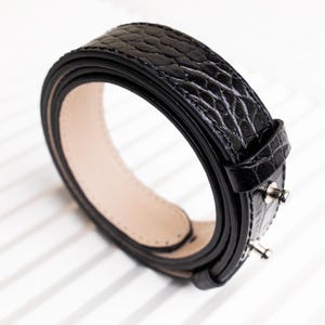 May include: A black leather belt with a crocodile embossed pattern. The belt is rolled up and the buckle is visible.