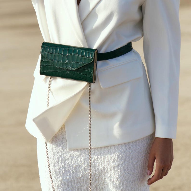 May include: A white blazer with a green crocodile-embossed leather fanny pack worn over a white skirt. The fanny pack has a silver chain strap.
