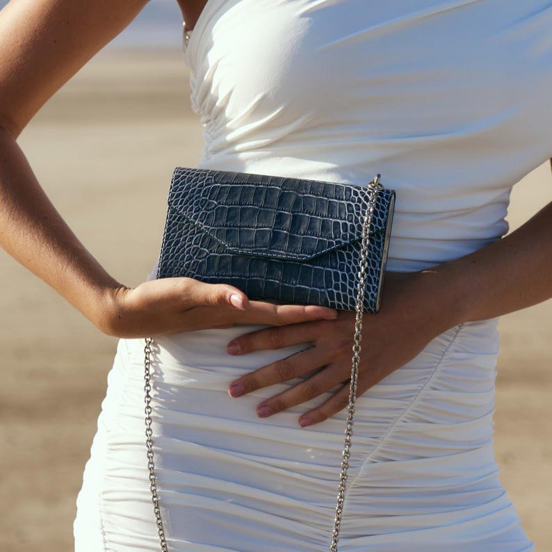 May include: A woman is holding a small, dark blue, crocodile-embossed clutch purse with a silver chain strap. The purse is closed and has a flap.