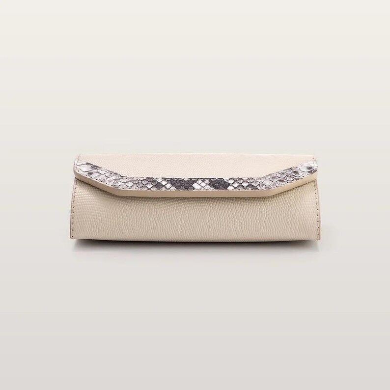 May include: A rectangular clutch bag in a light beige textured material. The front flap features a decorative band with a grey and white snakeskin pattern. The bag has a structured, elongated shape with visible stitching along the side edges.