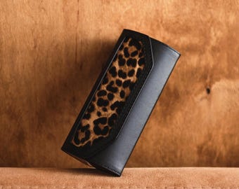 May include: A rectangular black wallet featuring a central panel with a brown and black leopard print pattern. The wallet has a smooth finish on the black sections and a textured appearance on the animal print section, with visible stitching.