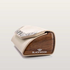 May include: A rectangular case with a dark brown wood grain side panel featuring the white text BLACKWOOD and a logo. The main body is a light cream color with a textured, wavy pattern and a flap closure accented with a metallic, scale-like detail.