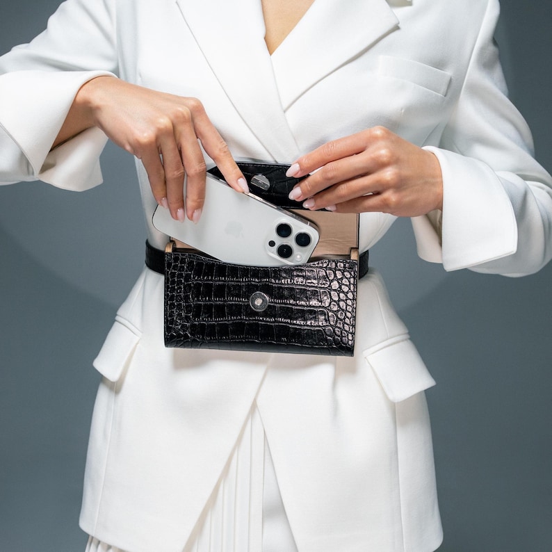 May include: A white blazer with a black crocodile-embossed leather belt bag. A white smartphone is partially visible inside the bag.