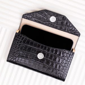 May include: Black leather clutch with a crocodile pattern and a snap closure. The clutch is open and a black smartphone is inside.