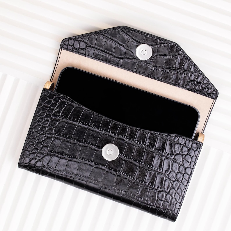 May include: Black leather clutch with a crocodile pattern and a snap closure. The clutch is open and a black smartphone is inside.