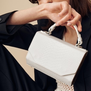 Puede incluir: Un bolso de mano de cuero blanco con un acabado texturizado y un cierre plateado. El bolso tiene un asa superior y una correa para el hombro desmontable.