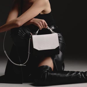 May include: A structured handbag featuring a white top flap with a textured snakeskin pattern and a solid black base. The bag includes a white top handle and a detachable, thin white shoulder strap with silver-toned metal hardware attachments.