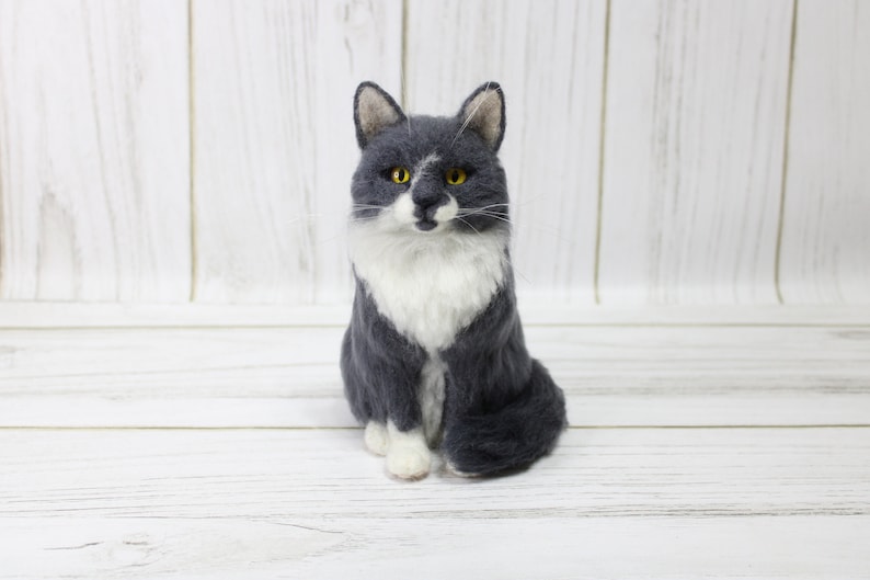 May include: A handcrafted felt cat figurine in a seated position. The cat has a gray and white coat, with yellow eyes and a white patch on its nose. The cat is sitting on a white wooden surface.