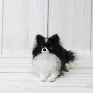 K&ouml;nnte beinhalten: Eine schwarz-wei&szlig;e Nadelfilz-Hundefigur, die auf einer wei&szlig;en Holzoberfl&auml;che liegt. Der Hund hat eine wei&szlig;e Brust und einen schwarzen Kopf mit schwarzen Ohren und einer wei&szlig;en Schnauze.