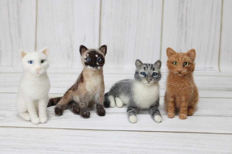May include: Four handcrafted felt cat figurines in various colors and patterns. A white cat with blue and green eyes, a Siamese cat, a gray tabby, and a ginger cat with green eyes are displayed. The cats are posed on a white wooden surface.
