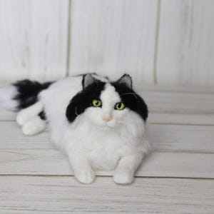 May include: A handmade, fluffy cat figurine in a relaxed pose. The cat has a white body with black markings on its head and back. It has green eyes and a long, fluffy tail. The cat is lying on a light-colored wooden surface.