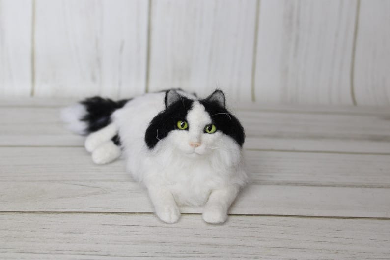 May include: A handmade, fluffy cat figurine in a relaxed pose. The cat has a white body with black markings on its head and back. It has green eyes and a long, fluffy tail. The cat is lying on a light-colored wooden surface.