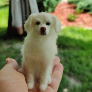 Puede incluir: Una figurita de perro de fieltro de aguja blanca. El perro está sentado con las patas metidas debajo de él. Tiene un pelaje blanco y esponjoso y ojos marrones.
