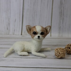 K&ouml;nnte beinhalten: Eine wei&szlig;e und braune Nadelfilz-Chihuahua-Hundefigur mit braunen Augen. Die Figur liegt auf einer wei&szlig;en Holzoberfl&auml;che mit zwei braunen Weidenkugeln.