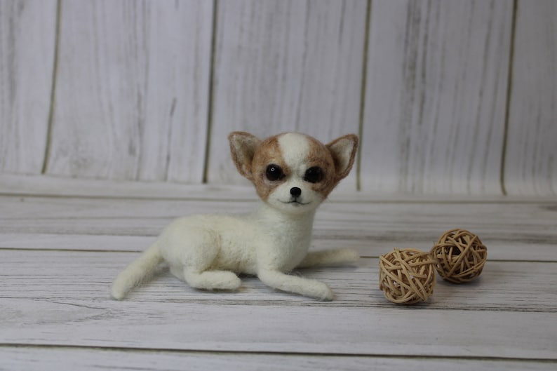 K&ouml;nnte beinhalten: Eine wei&szlig;e und braune Nadelfilz-Chihuahua-Hundefigur mit braunen Augen. Die Figur liegt auf einer wei&szlig;en Holzoberfl&auml;che mit zwei braunen Weidenkugeln.
