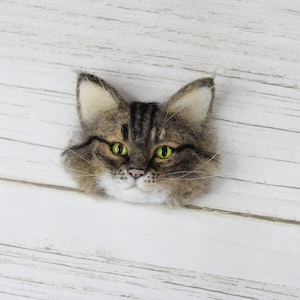 Puede incluir: Una broche de fieltro con forma de cabeza de gato con pelo marr&oacute;n y blanco, ojos verdes y nariz blanca. La broche est&aacute; sobre un fondo de madera blanco.