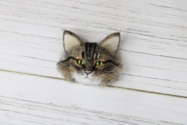 Puede incluir: Una broche de fieltro con forma de cabeza de gato con pelo marr&oacute;n y blanco, ojos verdes y nariz blanca. La broche est&aacute; sobre un fondo de madera blanco.