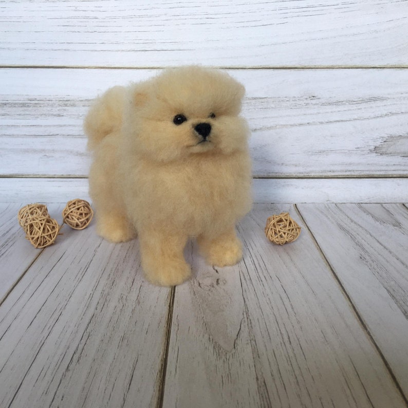 K&ouml;nnte beinhalten: Ein cremefarbenes, flauschiges, nadelgefilztes Pomeranian-Welpen-Spielzeug. Das Spielzeug steht auf einer hellen Holzoberfl&auml;che.