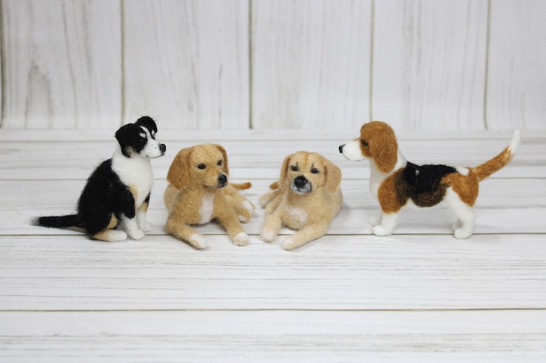 K&ouml;nnte beinhalten: Vier Nadelfilz-Hundefiguren. Ein schwarz-wei&szlig;er Hund sitzt links, ein hellbrauner Hund liegt links, ein hellbrauner Hund liegt rechts und ein braun-schwarz-wei&szlig;er Hund steht rechts.