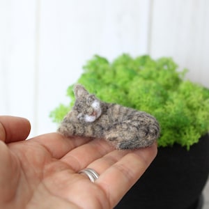 May include: A small, needle-felted figurine of a sleeping tabby cat with brown, grey, and white fur. The cat is curled in a resting position with its head tucked down, showcasing a textured, soft appearance created from wool fibres.