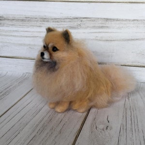 K&ouml;nnte beinhalten: Eine kleine, flauschige, cremefarbene, nadelgefilzte Pomeranian-Hundefigur. Der Hund sitzt mit dem Kopf zur Seite gedreht und dem Schwanz hinter sich eingerollt.
