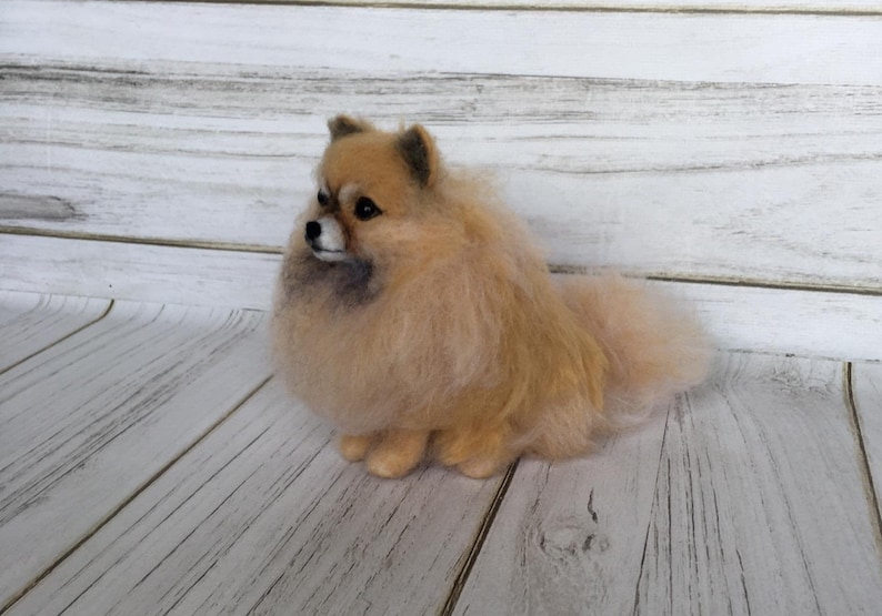 K&ouml;nnte beinhalten: Eine kleine, flauschige, cremefarbene, nadelgefilzte Pomeranian-Hundefigur. Der Hund sitzt mit dem Kopf zur Seite gedreht und dem Schwanz hinter sich eingerollt.