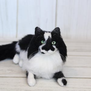 May include: A handmade, plush cat figurine in a lying pose. The cat has black and white fur with green eyes. The cat's fur is black on its head, back, and tail, with white fur on its chest, belly, and paws. The cat is lying on a light wood surface.