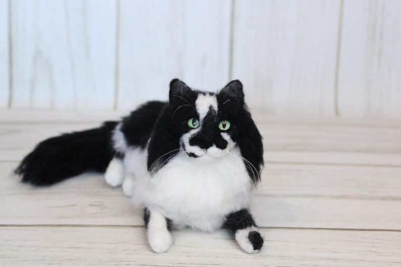 May include: A handmade, plush cat figurine in a lying pose. The cat has black and white fur with green eyes. The cat's fur is black on its head, back, and tail, with white fur on its chest, belly, and paws. The cat is lying on a light wood surface.