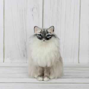 May include: A handcrafted, seated cat figurine with a fluffy, light-colored coat and striking blue eyes. The cat has gray markings on its face and ears, and its fur appears soft and detailed. The figurine is set against a white wooden background.
