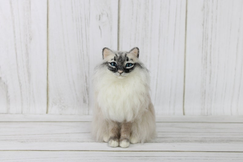 May include: A handcrafted, seated cat figurine with a fluffy, light-colored coat and striking blue eyes. The cat has gray markings on its face and ears, and its fur appears soft and detailed. The figurine is set against a white wooden background.