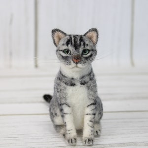 May include: A handcrafted gray and white cat figurine with green eyes and a pink nose. The cat is sitting upright, with its tail curled slightly. The figurine is made of felted wool and is a decorative item.