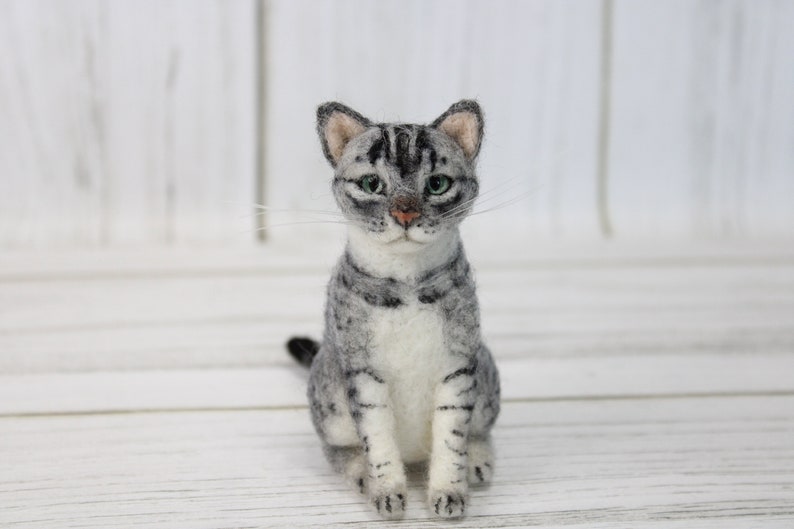 May include: A handcrafted gray and white cat figurine with green eyes and a pink nose. The cat is sitting upright, with its tail curled slightly. The figurine is made of felted wool and is a decorative item.