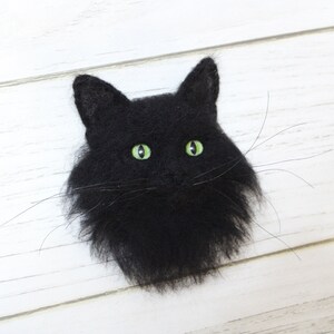 Puede incluir: Una cabeza de gato negra de fieltro con ojos verdes. El gato est&aacute; mirando hacia adelante y tiene un aspecto esponjoso y realista.