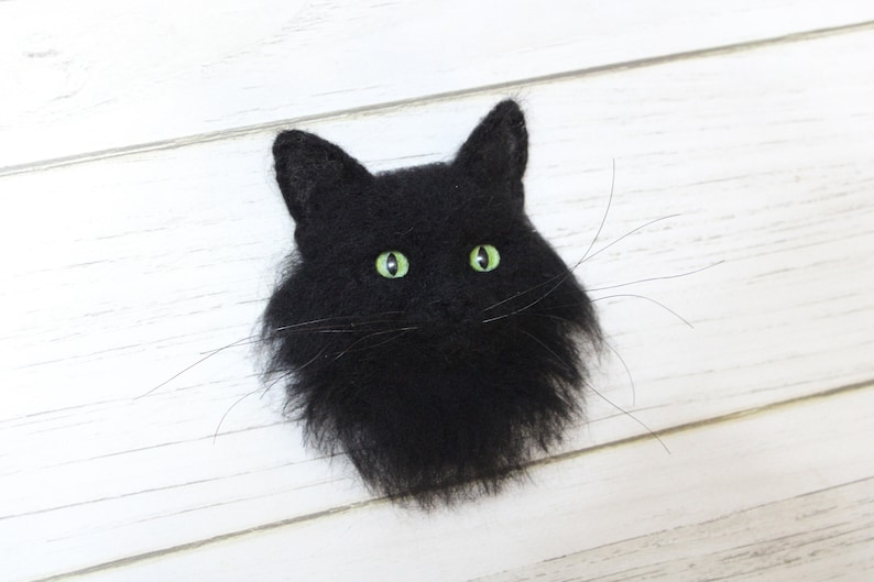 Puede incluir: Una cabeza de gato negra de fieltro con ojos verdes. El gato est&aacute; mirando hacia adelante y tiene un aspecto esponjoso y realista.